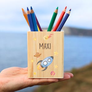 Könnte beinhalten: Ein Stifthalter aus Holz mit dem Namen "MAXI" auf der Vorderseite. Der Halter ist mit einer Rakete, Planeten und Sternen verziert. Er enthält mehrere Buntstifte.