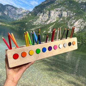 Pode incluir: Um porta-lápis de madeira com acabamento natural, cheio de lápis coloridos. O suporte tem uma fileira de círculos coloridos na frente. O fundo apresenta um lago e montanhas.