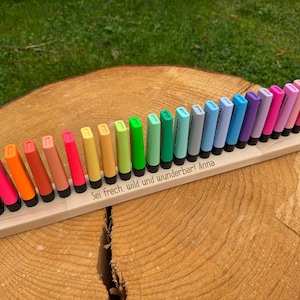 May include: A wooden organizer holding a collection of colorful highlighters. The highlighters are arranged in a row, showcasing a spectrum of vibrant colors. The base has the text "Sei frech, wild und wunderbar! Anna".