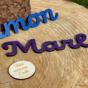 May include: Two wooden names, "Simon" in blue and "Marla" in purple, are displayed on a wooden surface. A small round wooden sign with the text "For unsere Erde" is also visible.