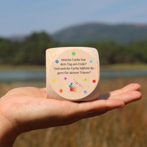 May include: A wooden cube with colourful dots and the text "Welche Farbe hat dein Tag am Ende? Und welche Farbe hättest du gern für deinen Traum?" is held in a hand. The cube has a painted image of a paintbrush and paint.
