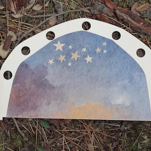 Op de afbeelding: Een houten, boogvormig decoratief stuk met een geschilderd aquarelontwerp. Het ontwerp toont een nachthemel met sterren en een kleurengradiënt van blauw, paars en geel. Het stuk heeft verschillende cirkelvormige uitsparingen.