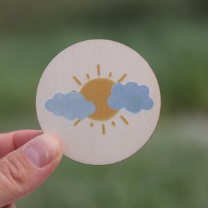 May include: A round wooden disc with a painted design of a yellow sun with rays and two blue clouds.
