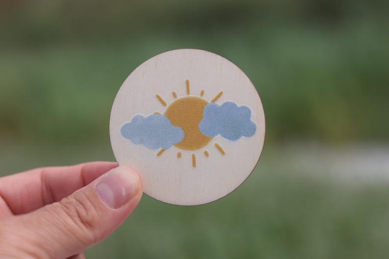 May include: A round wooden disc with a painted design of a yellow sun with rays and two blue clouds.