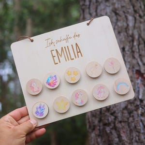 Könnte beinhalten: Eine Holztafel mit dem Namen "EMILIA" und dem Satz "Ich schaffe das" in Gold aufgedruckt. Sie verfügt über zehn kreisförmige Holzmarken mit Einhorn- und Regenbogenmotiven. Eine braune Lederschnur ist zum Aufhängen befestigt.