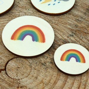 May include: Two round wooden discs with a rainbow design. The larger disc has a larger rainbow with red, orange, yellow, green, blue, indigo, and violet colors. The smaller disc has a smaller rainbow with the same colors.
