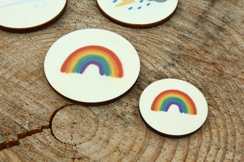 May include: Two round wooden discs with a rainbow design. The larger disc has a larger rainbow with red, orange, yellow, green, blue, indigo, and violet colors. The smaller disc has a smaller rainbow with the same colors.