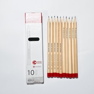 Könnte beinhalten: Ein Set aus zehn HB-Graphitstiften mit naturbelassenen hellen Holzgehäusen und rot getauchten Enden, angeordnet neben ihrer vertikalen weißen Pappschachtel. Die Schachtel trägt den Text SWISS WOOD, CARAN D'ACHE, SWISS MADE und die Zahl 10.