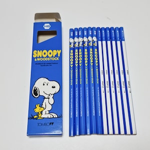 Op de afbeelding: Een set van 10 blauwe en witte potloden met Snoopy en Woodstock. De potloden zijn verpakt in een blauwe doos met de tekst "Snoopy & Woodstock" en "PEANUTS United Feature Syndicate, Inc."