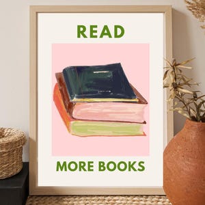 May include: A framed art print featuring a stack of three books with colorful covers against a pink background. The words "READ MORE BOOKS" are printed in green. The artwork is displayed on a neutral-toned wall.