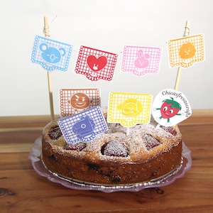 May include: A strawberry cake decorated with colourful paper flags featuring cartoon characters. The flags are attached to wooden skewers and are arranged in a circle around the cake. The cake is on a purple plate and the background is a light wood surface. The text "Chicasfresascrafts" is on a white circle with a red strawberry.