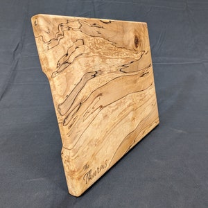 Könnte beinhalten: Ein großes, rechteckiges Schneidebrett aus hellem Holz mit einem einzigartigen Maserungsmuster. Das Brett hat abgerundete Kanten und ist mit dem Text "The Thurmans" graviert.