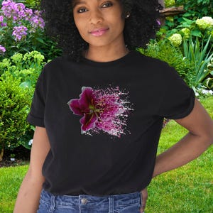 May include: A black short-sleeved t-shirt featuring a graphic print of a dark pink lily flower on the chest. The flower design transitions into a scattered, speckled pattern of pink and white dots, creating a vibrant contrast against the black fabric.