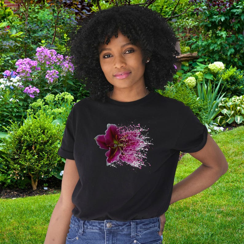 May include: A black short-sleeved t-shirt featuring a graphic print of a dark pink lily flower on the chest. The flower design transitions into a scattered, speckled pattern of pink and white dots, creating a vibrant contrast against the black fabric.