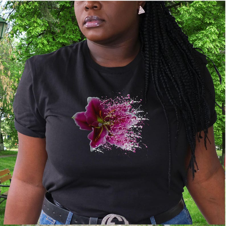 May include: A black short-sleeved t-shirt featuring a graphic print of a deep magenta lily flower on the chest. The floral design transitions into a white and magenta paint splatter effect that extends horizontally across the front of the garment.