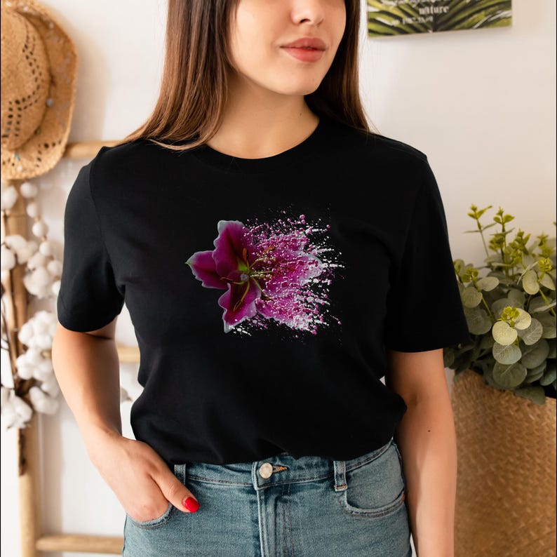 May include: A black short-sleeve t-shirt featuring a graphic print of a dark purple lily flower on the chest. The flower design transitions into a spray of white and pink paint splatters and glitter-like particles extending to the right side.