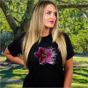 May include: A black short-sleeved t-shirt featuring a graphic print of a dark purple lily flower on the chest. The floral design includes a splatter effect with white and pink paint-like dots extending outward from the center of the bloom.