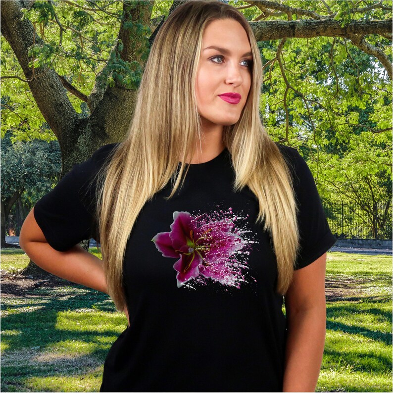 May include: A black short-sleeved t-shirt featuring a graphic print of a dark purple lily flower on the chest. The floral design includes a splatter effect with white and pink paint-like dots extending outward from the center of the bloom.