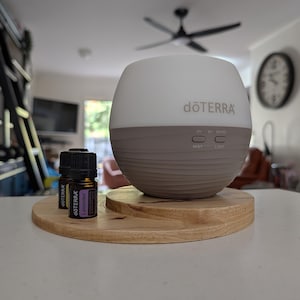 May include: A white and taupe essential oil diffuser with the brand name "dōTERRA" printed on the front. Two small essential oil bottles sit on a wooden tray. One bottle is labeled "Lavender".