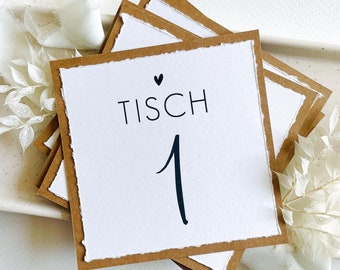 Könnte beinhalten: Eine weiße Karte mit braunem Rand und dem Text "Tisch 1" mit einem kleinen Herz über dem Text. Die Karte liegt auf anderen ähnlichen Karten.