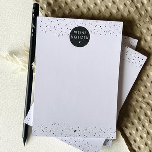 Könnte beinhalten: Ein Stapel weißer Notizblöcke mit schwarzem gepunkteten Rand und einem schwarzen Bleistift. Die Notizblöcke tragen den Text "MEINE NOTIZEN" in einem schwarzen Kreis. Der Bleistift ist schwarz mit weißem Text.