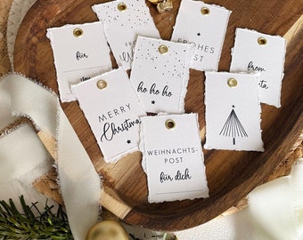May include: A collection of white gift tags with torn edges, each featuring gold eyelets. The tags display various festive messages in black script, including "Merry Christmas," "Ho Ho Ho," and "From Santa." The tags are arranged on a wooden tray.