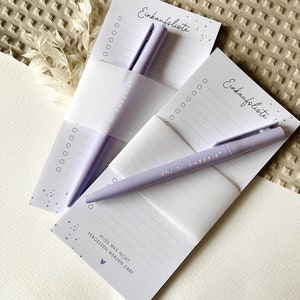 May include: Two purple shopping list pads with a purple pen on each. The pads have the text "Einkaufsliste" which means shopping list in German. The text "Alles was nicht vergessen werden darf" which means "Everything that shouldn't be forgotten" is on the bottom of one of the pads.
