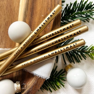 Könnte beinhalten: Goldfarbene Metallstrohhalme mit dem schwarzen Schriftzug "HAVE A MAGICAL CHRISTMAS". Die Strohhalme liegen auf einem Holzbrett mit weißen Weihnachtskugeln und grünen Tannenzweigen. Festliche Weihnachtsdekoration.