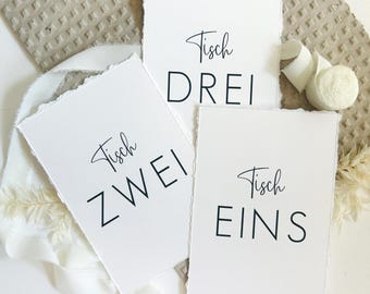 Könnte beinhalten: Drei rechteckige weiße Papierkarten mit Büttenrand und schwarzem Aufdruck. Die Karten zeigen die deutschen Wörter Tisch EINS, Tisch ZWEI und Tisch DREI in einer Kombination aus eleganter Schreibschrift und fetter serifenloser Typografie.