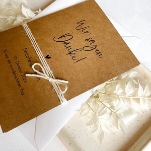 May include: A brown card with the text "Wir sagen Danke!" and "Nicole & ... 10. Oktober 2024 Es war ein wundersch&ouml;ner Tag."  The card is tied with twine and has a small heart drawn on it.