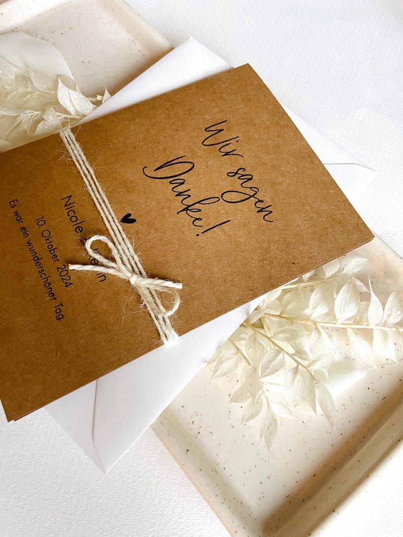 May include: A brown card with the text "Wir sagen Danke!" and "Nicole & ... 10. Oktober 2024 Es war ein wundersch&ouml;ner Tag."  The card is tied with twine and has a small heart drawn on it.