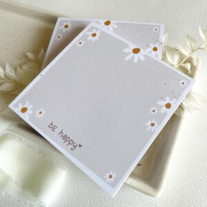 May include: Two gray square notepads with a white daisy border and the text "be happy" on the bottom. The notepads are on a speckled surface.