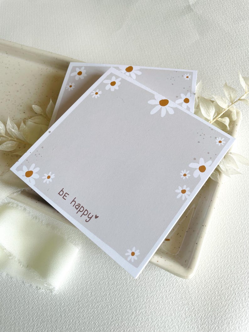 May include: Two gray square notepads with a white daisy border and the text "be happy" on the bottom. The notepads are on a speckled surface.