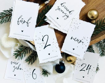 May include: White paper tags with black handwritten numbers and text, including "fünf", "2", "6", "neun", "vierzehn", "24", and "dreiund zwanzig". Each tag has a torn edge and a small heart. The tags are arranged with Christmas ornaments and greenery.