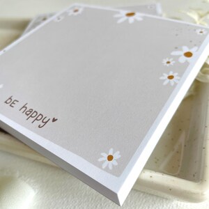 May include: A gray notepad with a white border and a white daisy pattern. The notepad has the text "be happy" written on it in brown.