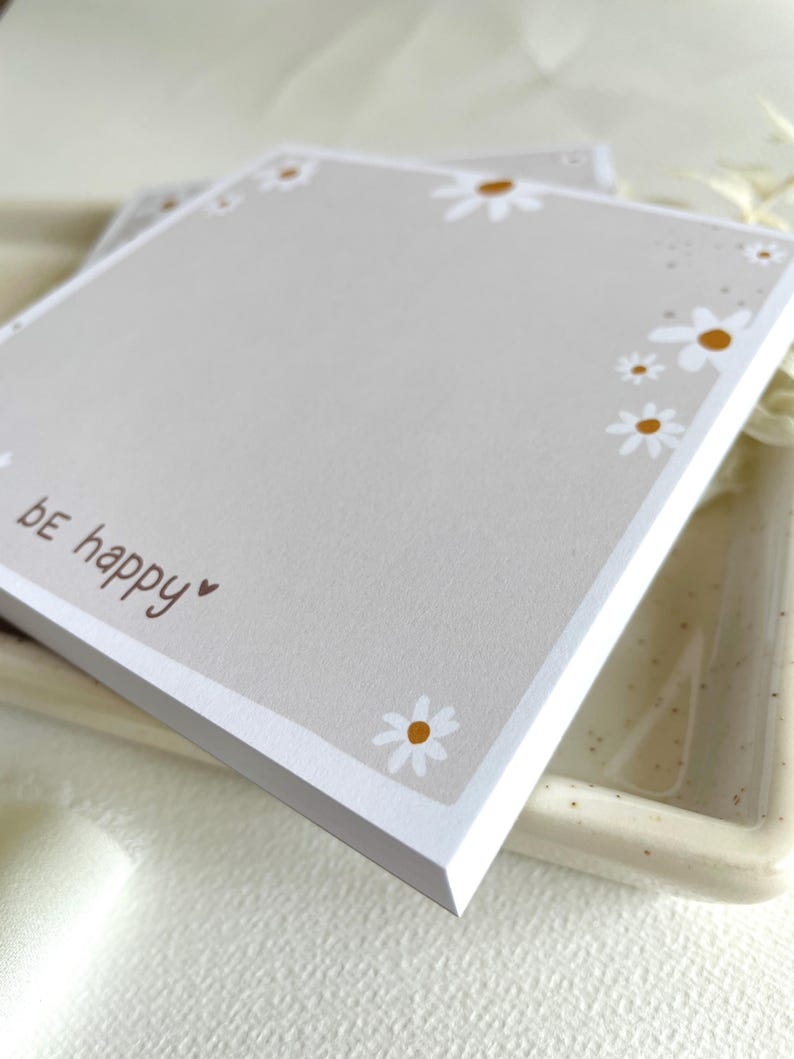 May include: A gray notepad with a white border and a white daisy pattern. The notepad has the text "be happy" written on it in brown.