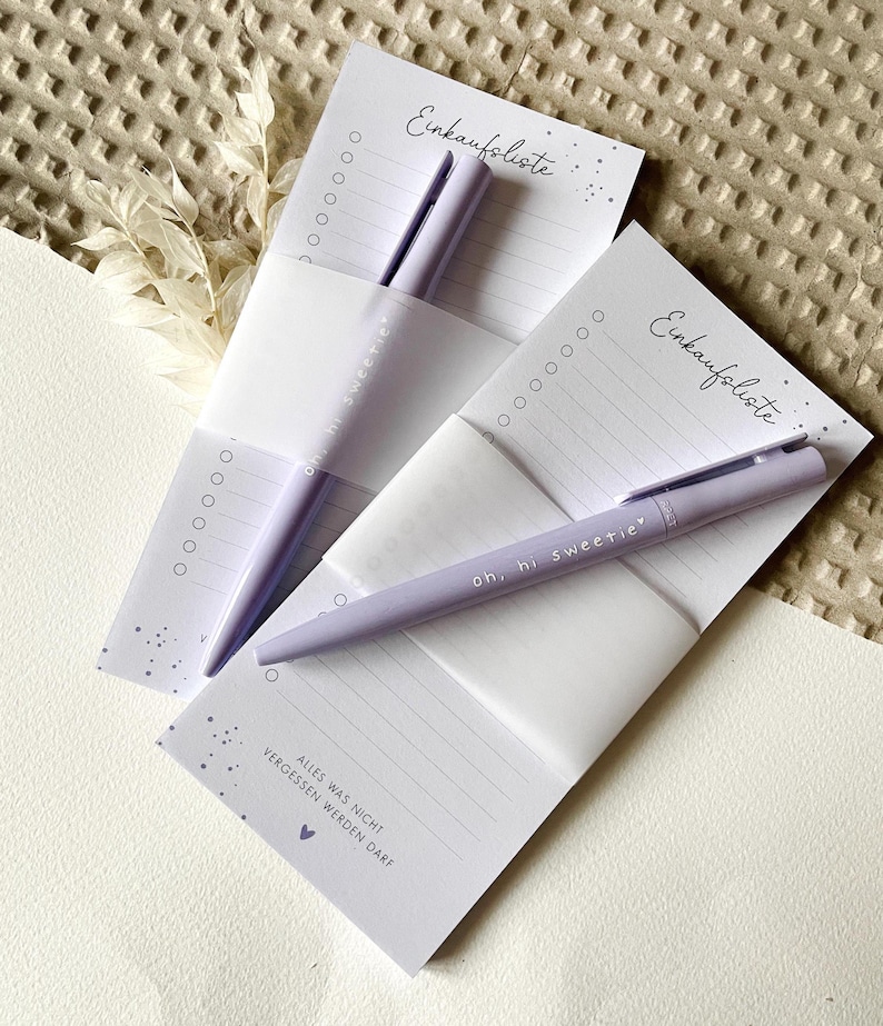 May include: Two purple shopping list pads with a purple pen on each. The pads have the text "Einkaufsliste" which means shopping list in German. The text "Alles was nicht vergessen werden darf" which means "Everything that shouldn't be forgotten" is on the bottom of one of the pads.