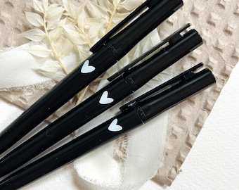 May include: Three black pens, each featuring a white heart detail. They are positioned diagonally against a textured, light brown backdrop, with white ribbon and dried flowers. The pens have a simple, elegant design.