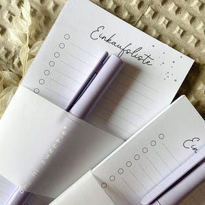 May include: Two purple notepad sets with a purple pen on each. The notepads have a white background with a dotted pattern and the text "Einkaufsliste" on the top. The pens have the text "ohni sweetie" on them.