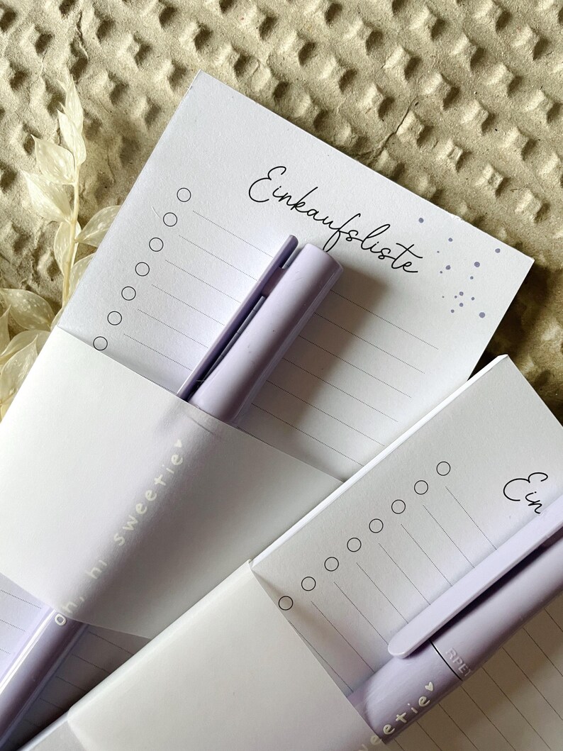 May include: Two purple notepad sets with a purple pen on each. The notepads have a white background with a dotted pattern and the text "Einkaufsliste" on the top. The pens have the text "ohni sweetie" on them.