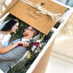 May include: A brown card with the text "Wir sagen Danke!" tied with twine and a small heart. The card is placed on top of a stack of photos of a bride and groom.