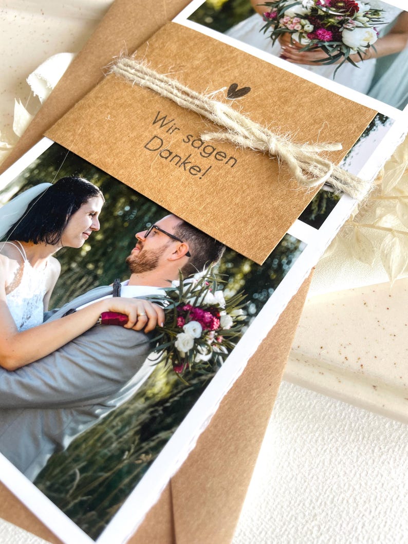 May include: A brown card with the text "Wir sagen Danke!" tied with twine and a small heart. The card is placed on top of a stack of photos of a bride and groom.
