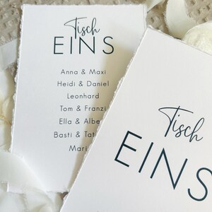 K&ouml;nnte beinhalten: Zwei wei&szlig;e rechteckige Karten mit B&uuml;ttenrand, die den schwarzen Text Tisch EINS und eine Namensliste mit Anna & Maxi, Heidi & Daniel, Leonhard, Tom & Franzi, Ella & Albert, Basti & Tatjana sowie Maria zeigen, akzentuiert durch ein wei&szlig;es Band.
