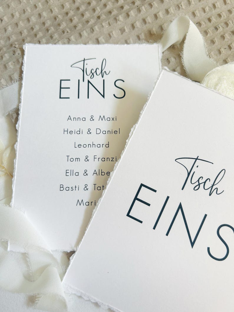 K&ouml;nnte beinhalten: Zwei wei&szlig;e rechteckige Karten mit B&uuml;ttenrand, die den schwarzen Text Tisch EINS und eine Namensliste mit Anna & Maxi, Heidi & Daniel, Leonhard, Tom & Franzi, Ella & Albert, Basti & Tatjana sowie Maria zeigen, akzentuiert durch ein wei&szlig;es Band.