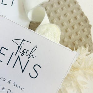 K&ouml;nnte beinhalten: Eine wei&szlig;e, rechteckige Karte mit B&uuml;ttenrand, die den schwarzen Schriftzug "Tisch EINS" sowie eine Namensliste mit "Anna & Maxi" und "& Daniel" zeigt. Die Karte ist mit einem wei&szlig;en Stoffband und getrockneten wei&szlig;en Pflanzenelementen verziert.