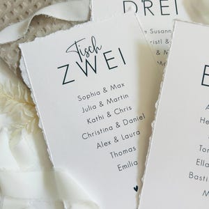 K&ouml;nnte beinhalten: Drei rechteckige wei&szlig;e Papierkarten mit B&uuml;ttenrand und schwarzem Aufdruck. Die Karten zeigen Tischnummern wie Tisch Zwei und Tisch Drei, gefolgt von Namenslisten in einer serifenlosen Schrift und einem kleinen schwarzen Herzsymbol.