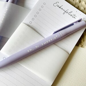 May include: A purple pen with the text "oh, hi sweetie" on it, lying on a white notepad with the text "Einkaufsliste" at the top. The notepad has lined paper and a few circles at the top.