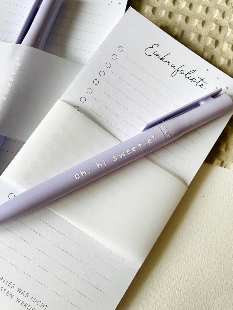 May include: A purple pen with the text "oh, hi sweetie" on it, lying on a white notepad with the text "Einkaufsliste" at the top. The notepad has lined paper and a few circles at the top.