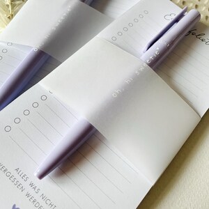 May include: Two purple pens are tucked into a white notepad with the text "oh, hi sweetie" and "Eu liste" on the covers. The notepad has lined pages and the text "ALLES WAS NICHT ERGESSEN WERDEN DARF" is printed on the bottom of the page.