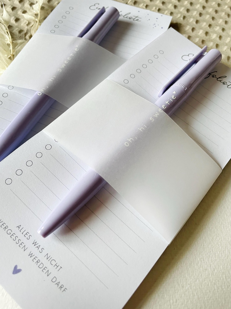 May include: Two purple pens are tucked into a white notepad with the text "oh, hi sweetie" and "Eu liste" on the covers. The notepad has lined pages and the text "ALLES WAS NICHT ERGESSEN WERDEN DARF" is printed on the bottom of the page.